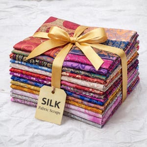 Könnte beinhalten: Ein Stapel bunter Seidenstoffreste, gebunden mit einem goldenen Band und einer Schleife. Die Stoffe zeigen verschiedene Muster und Farben, darunter Rot, Rosa, Lila und Orange. Ein Etikett mit der Aufschrift "SILK Fabric Scraps" hängt am Band.