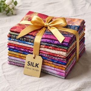 Pode incluir: Uma pilha de retalhos de tecido de seda dobrados em várias cores, incluindo vermelho, roxo, azul e dourado, com padrões tecidos intrincados. A pilha está amarrada com uma fita de cetim dourada e inclui uma etiqueta retangular com o texto SILK Fabric Scraps.