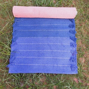 K&ouml;nnte beinhalten: Eine zusammengerollte rosa Yogamatte liegt auf einem blau- und indigogef&auml;rbten Webteppich mit wei&szlig;en Streifen. Der Teppich hat einen Fransenrand. Die Artikel werden auf gr&uuml;nem Gras pr&auml;sentiert.