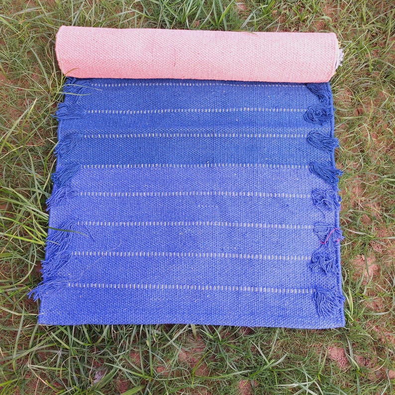 K&ouml;nnte beinhalten: Eine zusammengerollte rosa Yogamatte liegt auf einem blau- und indigogef&auml;rbten Webteppich mit wei&szlig;en Streifen. Der Teppich hat einen Fransenrand. Die Artikel werden auf gr&uuml;nem Gras pr&auml;sentiert.