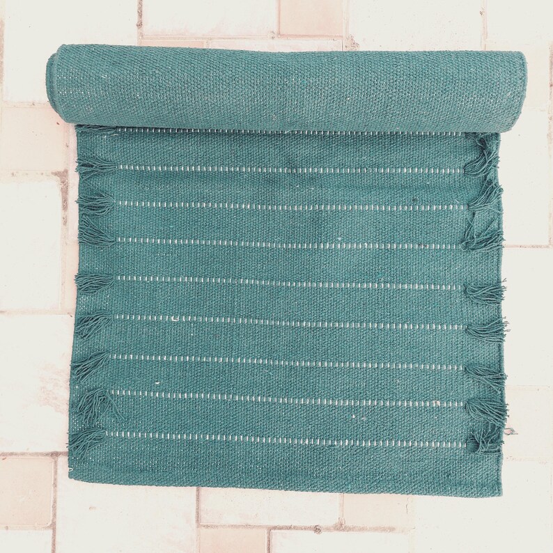 K&ouml;nnte beinhalten: T&uuml;rkisfarbener gewebter Teppich mit wei&szlig;en horizontalen Streifen und Fransen. Der Teppich ist teilweise aufgerollt und zeigt seine Textur und sein Design. Hergestellt aus strapazierf&auml;higem Material, geeignet f&uuml;r verschiedene R&auml;ume.