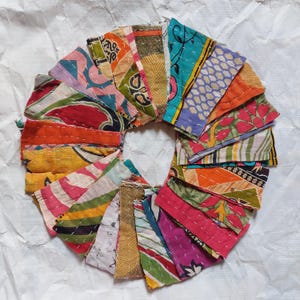 May include: A circular arrangement of colourful fabric pieces, each with unique patterns and colours, including pink, orange, green, and blue. The pieces are stitched together with white thread, creating a patchwork design. The background is a white, textured surface.