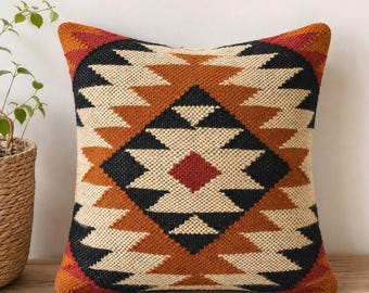 May include: Square decorative pillow with a geometric pattern in shades of black, tan, orange, and red. The pillow has a woven texture and a diamond-shaped design in the center. The pillow is placed on a wooden surface.