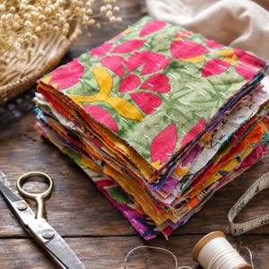 May include: A stack of colorful fabric squares with floral patterns, featuring pink, yellow, and green designs. The squares are hand-stitched with white thread. Scissors, a measuring tape, and thread are on a wooden surface.