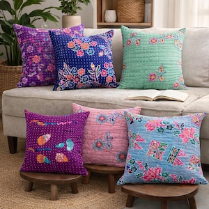 May include: A collection of decorative throw pillows in various colors and floral patterns. Pillows include purple, blue, pink, and green hues. The pillows are square-shaped and appear to be made of a quilted fabric. Some pillows are on a beige sofa, and others are on small wooden stools.
