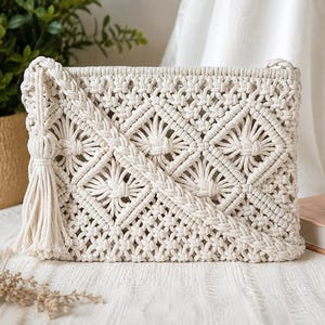 May include: A rectangular off-white macrame clutch bag featuring an intricate knotted diamond pattern, a braided strap, and a long decorative tassel attached to the side. The bag is constructed from thick cotton cord with a textured, woven finish.