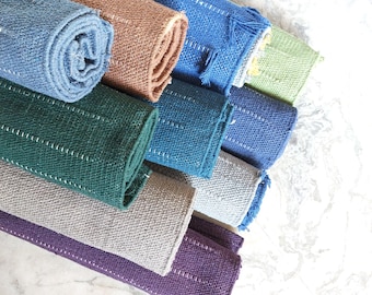 May include: A collection of rolled fabric placemats in various solid colors including blue, brown, green, gray, and purple. Each placemat features a textured weave and a series of parallel white stitched lines running along the length of the fabric.