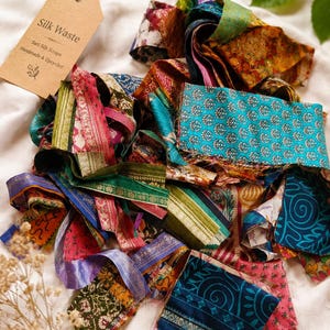 May include: A collection of multicoloured sari silk scraps featuring various intricate patterns and textures. A brown paper tag is attached with string, printed with the text Silk Waste, Sari Silk Scraps, and Handmade & Upcycled.