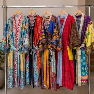 May include: Five long-sleeved robes hanging on a metal rack. Each robe features a unique patchwork design with vibrant colors including blue, red, yellow, and purple, finished with a matching fabric tie belt at the waist and wide sleeves.