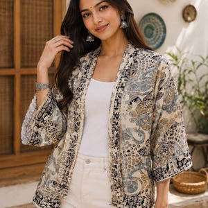 May include: A cream-colored open-front kimono jacket featuring a detailed black, blue, and tan floral and paisley pattern. The garment has three-quarter length sleeves and a straight hem, worn over a plain white tank top and cream-colored pants.