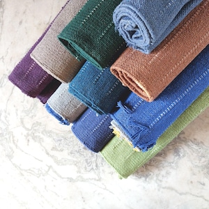 K&ouml;nnte beinhalten: Eine Sammlung farbenfroher, gewebter Stoffst&uuml;cke in Lila, Beige, Gr&uuml;n, Blau und Braun. Jedes St&uuml;ck ist rechteckig mit einer gen&auml;hten Bord&uuml;re, einige haben dekorative Fransen. Die Stoffe sind ordentlich angeordnet und zeigen ihre Texturen.