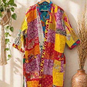 May include: A patchwork kimono robe in vibrant colors, including yellow, red, and purple. The robe features a mix of patterned fabrics and a contrasting teal lining. It hangs on a wooden hanger against a neutral wall.