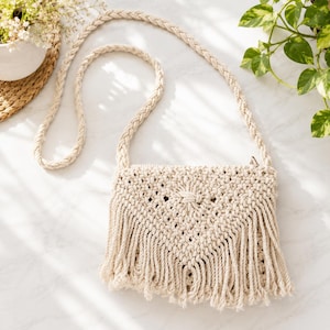 Può includere: Una borsa a tracolla in macramè color crema caratterizzata da una patta frontale a forma di V con intricati motivi a nodi e lunghe frange lungo il bordo inferiore. La borsa include una lunga tracolla intrecciata dello stesso materiale.