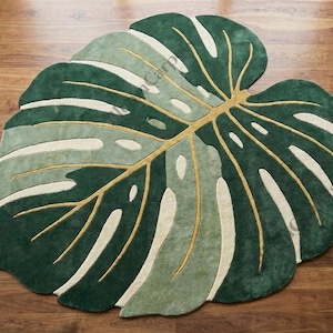 May include: A green and white leaf-shaped rug with a gold-colored vein design. The rug is made of a soft material and is placed on a wooden floor. The rug's design is inspired by a Monstera plant.