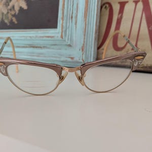 May include: Vintage cat-eye eyeglasses with a gold and brown frame. The lenses are clear, and the frame has decorative accents on the upper corners. The glasses are set against a blurred background.