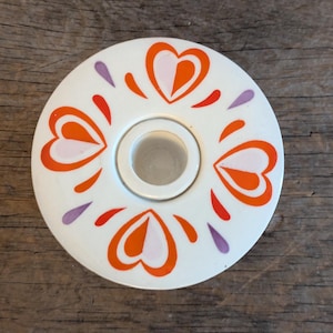 May include: White ceramic candle holder with a central opening and a decorative pattern of orange and pink hearts and purple teardrop shapes. The candle holder is set against a wooden background.