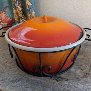 May include: Orange and yellow enamelware casserole dish with a white rim and a black metal stand. The dish has a gradient of color from orange to yellow.