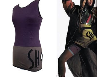 Puede incluir: Un vestido morado sin mangas con una sección de falda gris. La palabra "SHO" está impresa en letras negras grandes en la falda. El vestido tiene un escote redondo y una silueta ajustada. La imagen también muestra a una persona con una chaqueta con capucha y pantalones cortos.
