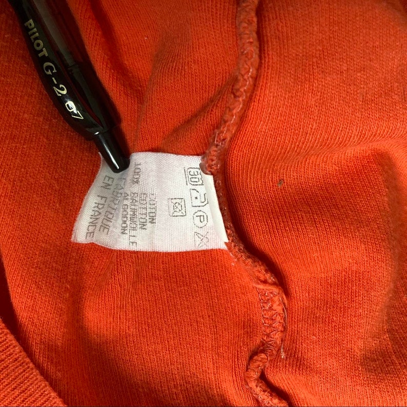 May include: White tag with black text and symbols. The text reads "100% COTTON", "FABRIQU&Eacute; EN ALGODON", "MADE IN LONDON", "FABRIQU&Eacute; EN FRANCE". The tag also has a symbol for 30 degrees Celsius washing, a symbol for non-chlorine bleach, a symbol for tumble drying at low temperature, and a symbol for ironing at low temperature.