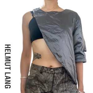 May include: A gray asymmetrical top with a black under layer and a zipper detail on the sleeve. The top is made of a shiny, silky fabric. The model is wearing the top with camouflage pants.