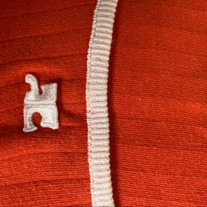 May include: A close-up of a red fabric with a white trim and a white embroidered logo of a stylized letter "H".