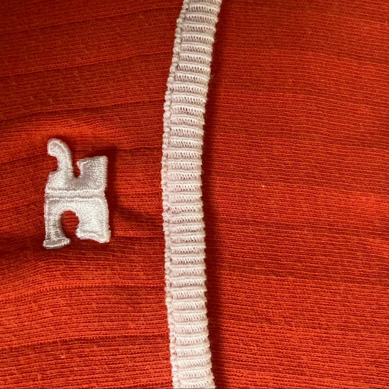 May include: A close-up of a red fabric with a white trim and a white embroidered logo of a stylized letter "H".