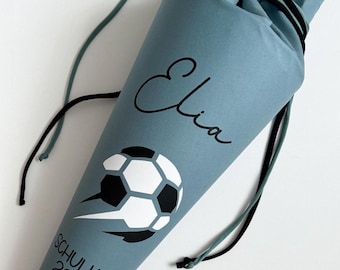 Peut inclure: Un sac en forme de cône bleu-vert avec un graphisme de ballon de football et le nom "Elia" en écriture noire. Le texte "SCHULKIND 2025" est également imprimé sur le sac. Il possède des cordons noirs et verts.
