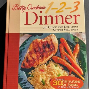 May include: A cookbook titled "Betty Crocker's 1-2-3 Dinner" with a photo of a cooked chicken breast, couscous, and carrots. The cover features the title in large red and orange letters, with the text "350 Quick and Delicious Supper Solutions."