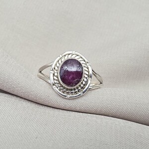 Peut inclure: Une bague en argent ornée d'une pierre précieuse ovale, d'un violet profond. La pierre est sertie dans un cadre décoratif en forme de corde, à l'intérieur d'un cadre ovale plus grand. L'anneau se divise en deux brins délicats de chaque côté.