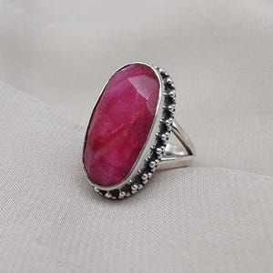 May include: A sterling silver ring featuring a large, oval-cut ruby gemstone. The ruby is a vibrant shade of red. The ring has a detailed, beaded border around the gemstone and a simple band. The ring is set against a neutral background.