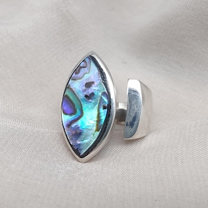 May include: A silver ring with an abalone shell marquise-shaped setting. The shell's iridescent surface shows blue, green, and purple hues. The ring has a contemporary design with a wide band.