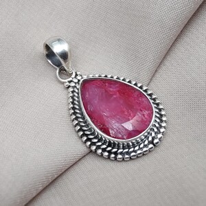 May include: A teardrop-shaped pendant featuring a faceted, vibrant red gemstone. The stone is set in a silver-toned, beaded frame with a matching bail for a necklace. The pendant is displayed on a neutral fabric background.