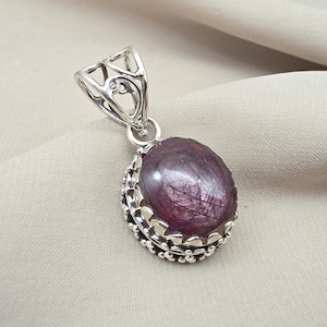 May include: A silver pendant with an oval, purplish-pink gemstone. The gemstone is set in a decorative, scalloped bezel. The bail is a detailed, openwork design. The pendant is displayed on a neutral-colored fabric.
