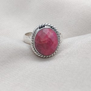 Puede incluir: Un anillo de plata con una piedra preciosa ovalada, facetada, de color rubí. La piedra preciosa está engastada en un borde retorcido en forma de cuerda. La banda del anillo es de plata pulida simple.