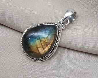 May include: A teardrop-shaped pendant with a polished labradorite stone, showcasing iridescent blues, greens, and yellows. The stone is set in a silver-toned metal frame with a rope-like border and a matching bail for a necklace.