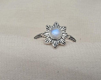 May include: A silver ring with a flower-shaped design, featuring a central, round, pale blue gemstone. The ring's band has a textured pattern. The flower petals are detailed and pointed. Jewellery.