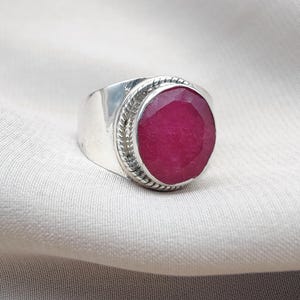 Peut inclure: Une bague en argent avec une grosse pierre précieuse rouge foncé de forme ovale. La pierre est sertie dans un anneau en argent poli avec une bordure texturée. La bague est présentée sur un fond en tissu neutre.