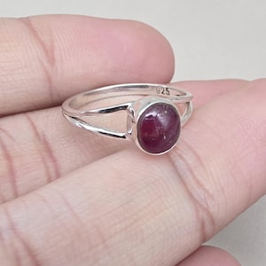 Könnte beinhalten: Ein Silberring mit geteiltem Schaft-Design. Der Ring hat einen großen, oval geformten, tiefroten Edelstein. Das Band ist mit "925" gestempelt. Dieser Ring ist ein Schmuckstück.