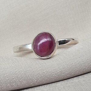 Puede incluir: Un anillo de plata con una gran piedra de rubí redonda con corte cabujón. El rubí es de un color rojo intenso, engastado en una banda de plata pulida y sencilla. El anillo se muestra sobre una tela de tono neutro.
