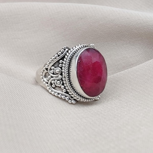 May include: A silver ring featuring an oval, deep red gemstone. The ring has an ornate, detailed design around the gemstone and on the band, with a textured, antique finish. The gemstone is the focal point, set in a bezel setting.