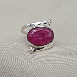 Könnte beinhalten: Ein silberner Ring mit einem großen, oval geschliffenen, tiefrosa Edelstein. Das Ringband umschlingt den Finger mit dekorativen Akzenten an den Enden. Der Edelstein ist der Blickfang und zeigt seine facettierte Oberfläche.