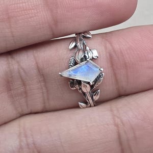 Op de afbeelding: Een zilveren ring met een blad- en wijnstokpatroon. Het heeft een rechthoekige, iriserende edelsteen met blauwe en witte tinten. De ring wordt vastgehouden door de vingers van een persoon.