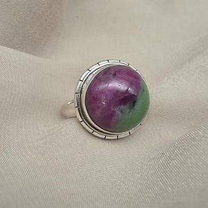 Puede incluir: Un anillo de plata con una piedra preciosa grande y redonda. La piedra preciosa presenta una mezcla de tonos magenta oscuro y verde. El anillo tiene una banda de plata texturizada y detallada alrededor de la piedra.