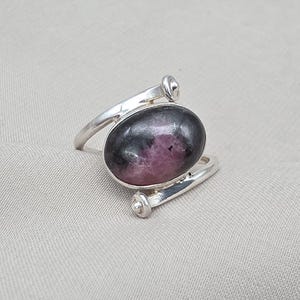 Könnte beinhalten: Ein silberner Ring mit einem großen, oval geformten Edelstein. Der Edelstein hat eine dunkelgraue und rosa Farbe. Das Band des Rings umschließt den Finger mit dekorativen silbernen Akzenten. Der Ring ist vor einem neutralen Hintergrund platziert.