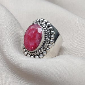 Puede incluir: Un anillo de plata con una gema grande, ovalada y de color rojo brillante. La gema está engastada en un bisel decorativo con un diseño de cuerda trenzada y pequeñas cuentas de plata. El anillo se muestra sobre una tela clara.