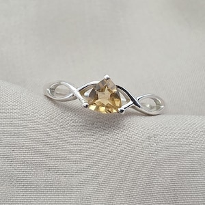 May include: A silver ring featuring a triangular, light-brown gemstone. The ring's band has an infinity symbol design on each side of the gemstone, creating a unique and elegant look. The gemstone is securely set in a prong setting.