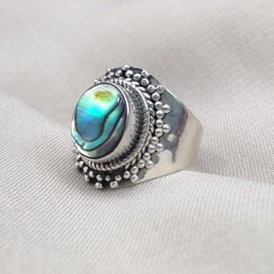 May include: A silver ring with an oval abalone shell displaying iridescent blues, greens, and golds. The ring has a detailed, textured setting with a rope-like border and small beaded accents, giving it a vintage or bohemian appearance.