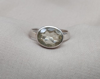 Könnte beinhalten: Ein Silberring mit einem großen, oval geschliffenen, hellgrünen Edelstein. Der Edelstein ist der Mittelpunkt, gefasst in einem schlichten, polierten Silberband. Der Ring wird auf einem neutralfarbenen Stoff präsentiert, der sein Design hervorhebt.