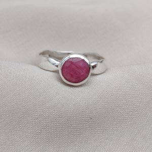 Op de afbeelding: Een zilveren ring met een ronde, gefacetteerde robijn edelsteen. De ringband heeft een eenvoudig, elegant ontwerp. De robijn heeft een levendige rode tint, gezet in een gepolijste zilveren bezel.