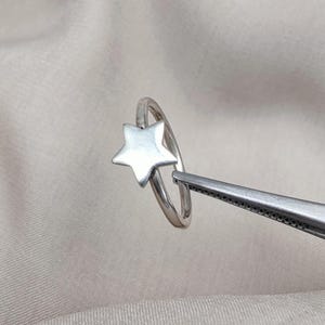 Op de afbeelding: Een zilveren ring met een stervormig ontwerp. De ring wordt door een pincet tegen een effen achtergrond gehouden. De ster is het belangrijkste kenmerk en presenteert een minimalistische en hemelse esthetiek.